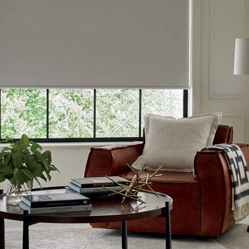 Designer roller shades covering three-fourths of a living room window with a comfy chair and coffee table in front