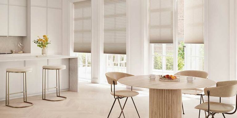 Duette® Honeycomb Shades covering floor to ceiling windows in a neutral kitchen. To the right of the photo is two chairs and a modern circular table