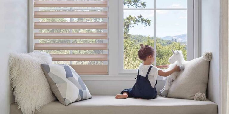 A little girl sitting in a built in window seat. She is looking out of windows that have cordless window coverings. She has stuffed animals and pillows around her as she peers outside on a sunny day.